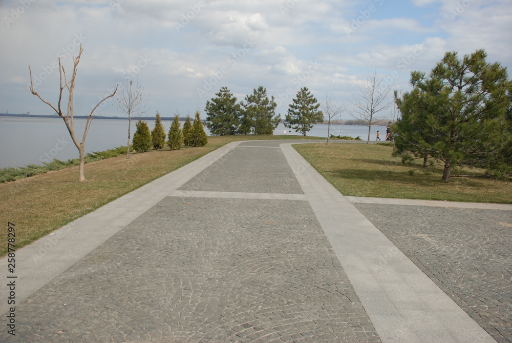A gray granite walkway runs in a park along the river. Around grow trees and shrubs.