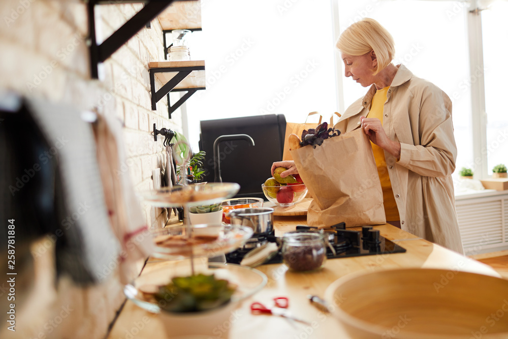 Content beautiful mature woman with short blond hair standing at kitchen counter and putting fruits in bowl while unpacking grocery bag - obrazy, fototapety, plakaty 