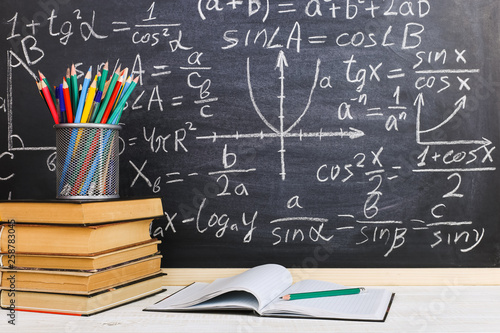 School desk in classroom, with books on background of chalk board with written formulas. Soncept Teacher's Day.