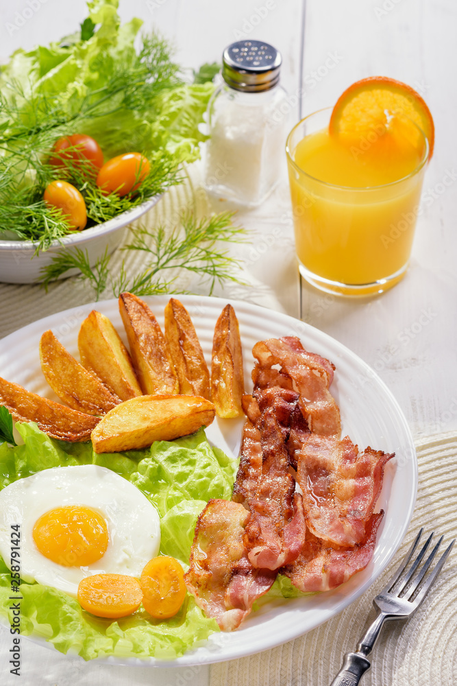 English breakfast-plate with fried bacon, with French fries, fried egg stands on a white table and a glass of fresh juice.