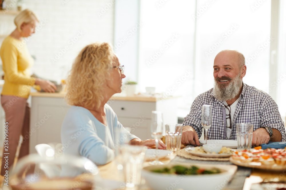 Jolly excited mature friends in casual clothing sitting at table and talking to each other while waiting for dinner, woman cooking in background Jolly excited mature friends in casual clothing sitting at table and talking to each other while waiting for dinner, woman cooking in background