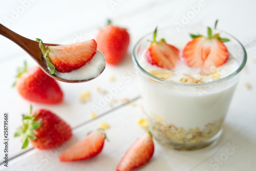 Yoghurt with fresh strawberries on wooden spoon. Decorated with some pieces of strawberries and muesli yoghurt in glass in background. Fresh food for breakfast concept.