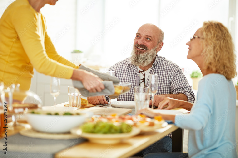 Lady in oven mitts brining hot dish and putting on wooden board, excited mature friends in anticipation of delicious dinner Lady in oven mitts brining hot dish and putting on wooden board, excited mature friends in anticipation of delicious dinner