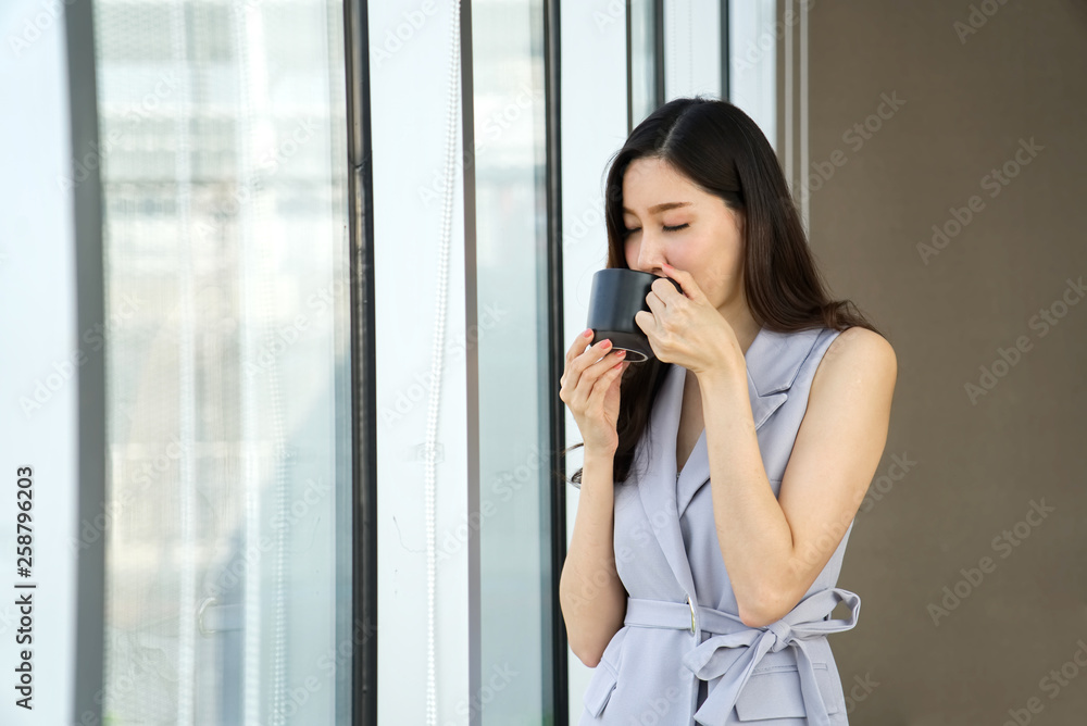 Asian smart beautiful girl talking a rest by drinking cup of coffee standing near the window in the office. She is closing her eyes and smelling the nice coffee. Smart working concept. Asian smart beautiful girl talking a rest by drinking cup of coffee standing near the window in the office. She is closing her eyes and smelling the nice coffee. Smart working concept.