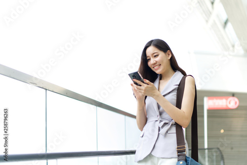 Asian office girl playing mobile phone standing on elevator in shopping mall. She is smiling and enjoy using the smartphone in her lifestyle. Technology and lifestyle concept.