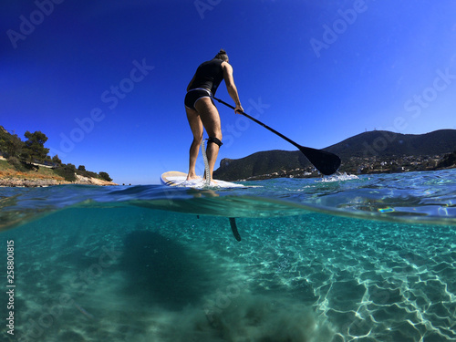 Underwater sea level photo of unidentified man on a SUP or Stand Up Paddle in turquoise crystal clear exotic mediterranean sandy beach