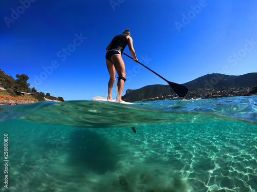 Underwater sea level photo of unidentified man on a SUP or Stand Up Paddle in turquoise crystal clear exotic mediterranean sandy beach