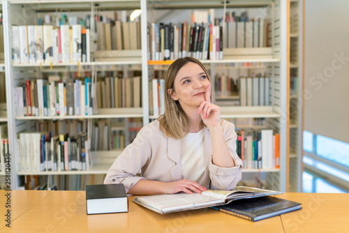 Chica joven hermosa caucásica con el dedo en la cara pensando contenta en la biblioteca con libros en la mesa