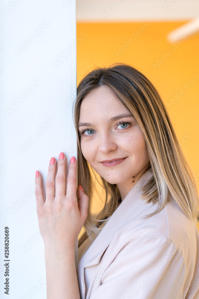Portrait of a beautiful young caucasian girl smiling with blue eyes leaning against a white wall with an orange background Portrait of a beautiful young caucasian girl smiling with blue eyes leaning against a white wall with an orange background