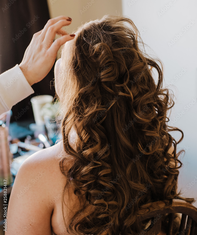 Naklejka premium Backstage work of professional hairdresser or make up artist. Close up hand touching up. Back view of woman with long beautiful curly hair natural colour.