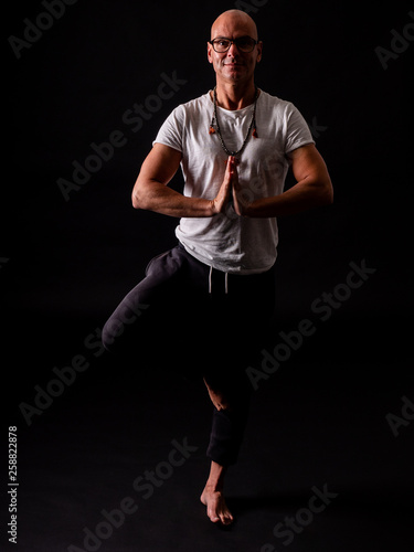 Buddhist monk practicing yoga on black background in long pants and glasses © Supermelon