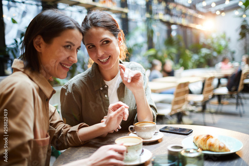Cheerful young woman in casual shirts sitting at table and drinking coffee while gossiping about people in cafe