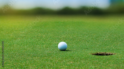 White Golf ball on green course near hole on blurred landscape of golf course in bright day time with copy space. Sport, Recreation, Relax in holiday concept