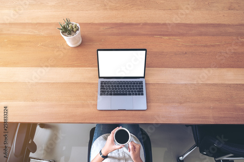 Top view mockup image of a woman using laptop with blank white desktop screen while drinking coffee on wooden table in office