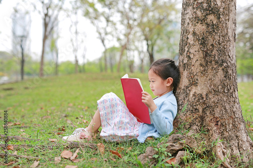 Cute little girl reading book in summer park outdoor lean against tree trunk in the summer garden.
