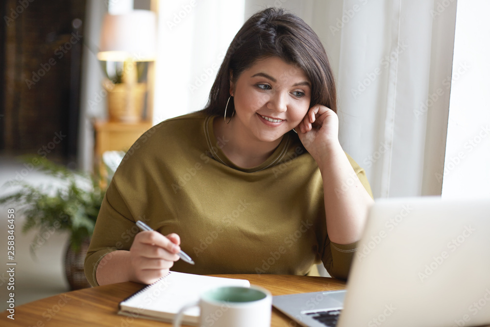 Indoor image of cheerful overweight student girl working on course paper, usign wireless internet connection on generic portable computer, making notes in copybook and smiling. Gadgets and education Indoor image of cheerful overweight student girl working on course paper, usign wireless internet connection on generic portable computer, making notes in copybook and smiling. Gadgets and education
