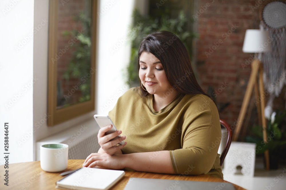 Overweight brunette female in xxl sweatshirt holding mobile phone typing text message via online messenger while working remotely from cozy cafe using laptop, writing down in copybook, having coffee