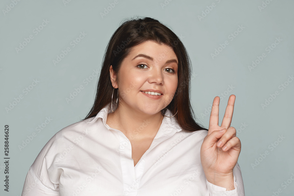 Portrait stylish happy confident young woman with curvy body showing peace sign, making victory gestures using middle and index fingers, smiling cheerfully at camera. Feelings, symbols and attitude Portrait stylish happy confident young woman with curvy body showing peace sign, making victory gestures using middle and index fingers, smiling cheerfully at camera. Feelings, symbols and attitude