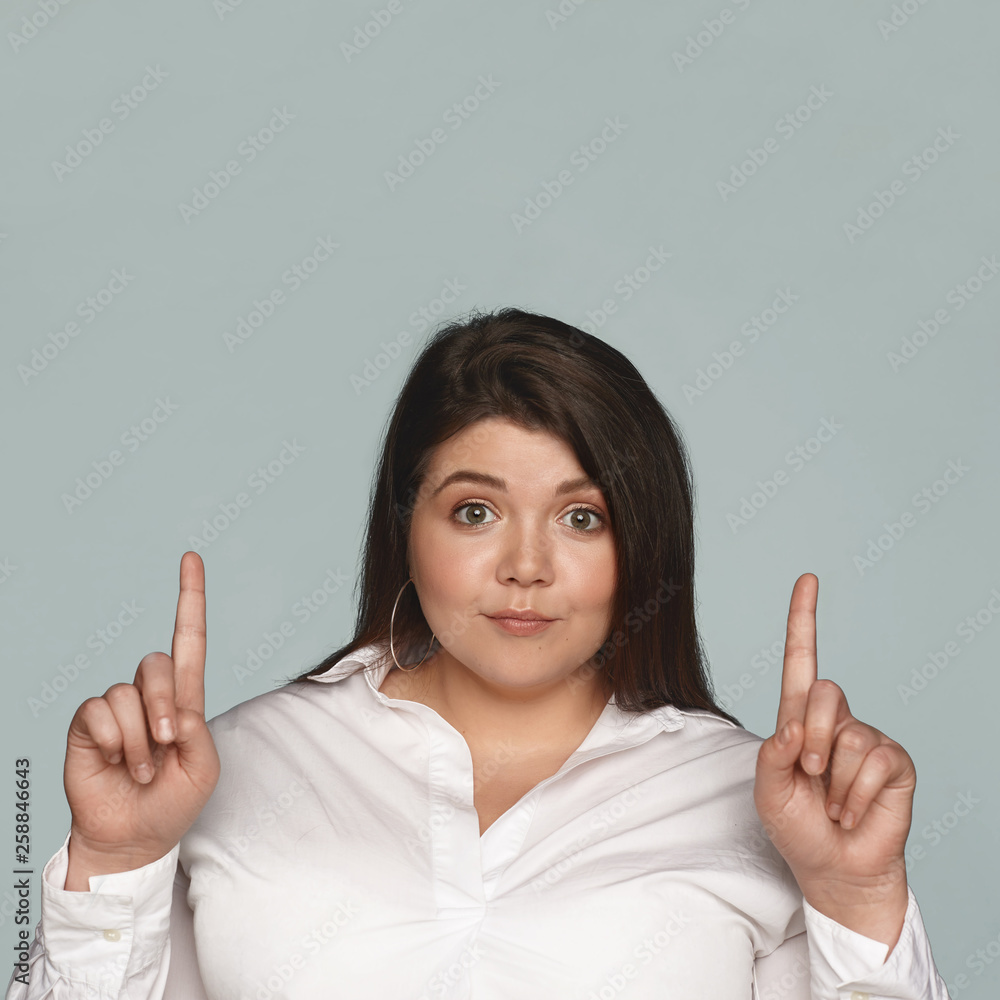 Happy cheerful young dark haired female with chubby cheeks and curvy large  body staring at camera with excited look, pointing both fore fingers  upwards, showing blank copyspace studio wall Stock Photo |