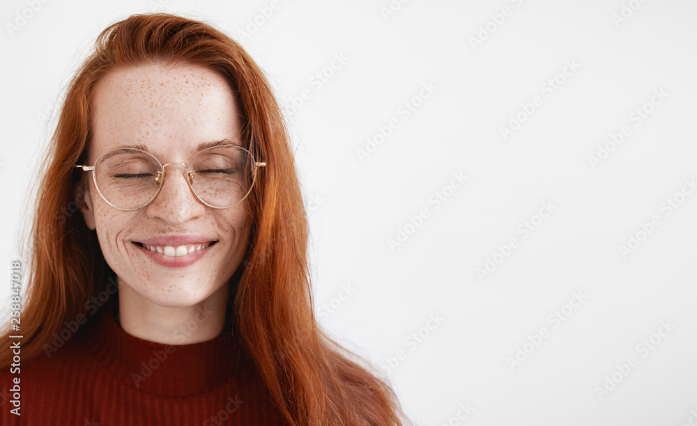 Joy and happiness concept. Isolated shot of beautiful young redhead freckled female in trendy eyewear, closing eyes, smiling with great pleasure, imagining or recollecting something pleasant