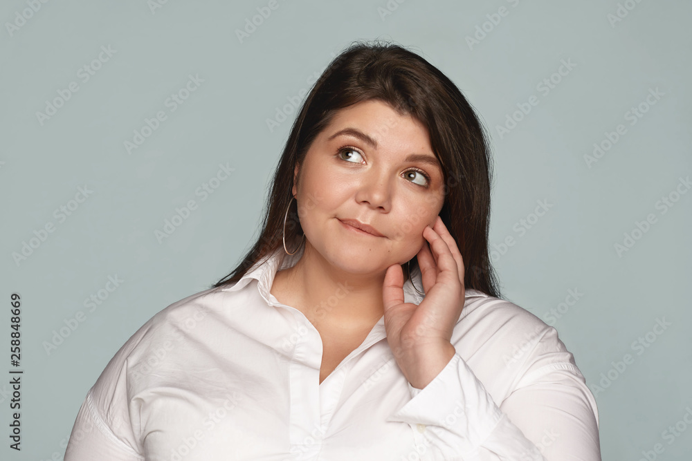Portrait of beautiful overweight young female employee wearing white shirt and round earrings looking up and biting lip, having pensive expression, thinking what healthy food eat for lunch Portrait of beautiful overweight young female employee wearing white shirt and round earrings looking up and biting lip, having pensive expression, thinking what healthy food eat for lunch