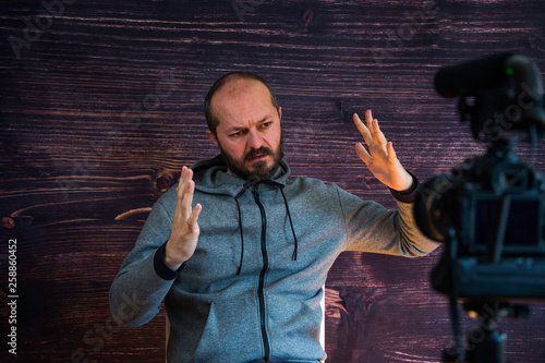 Bearded middle age man in hoody sitting and talking in front of DSLR camera, video blogging concept