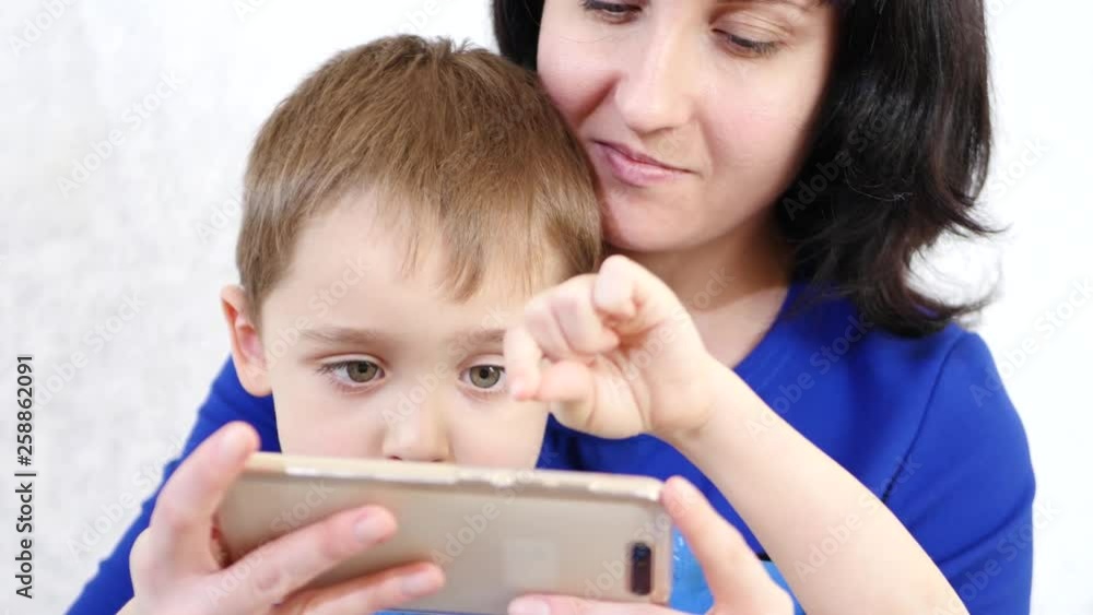 A woman holds a little boy hugging him. The child touches the touch screen of the phone and plays a child s game on a smart phone. Life is online. Education and development. A woman holds a little boy hugging him. The child touches the touch screen of the phone and plays a child s game on a smart phone. Life is online. Education and development.