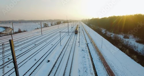 Drone flying backwards, capturing a train slowly approaching a staition during the winter, surrounded by fresh fallen snow, near a bicycle road. Between a dense green forest and empty fields. photo