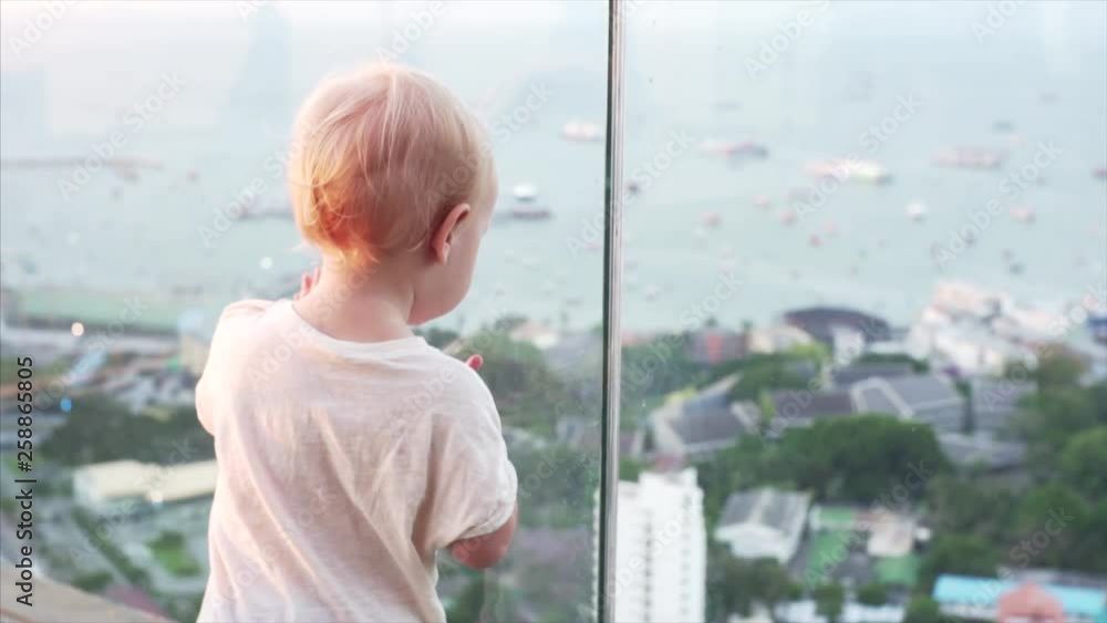 Close up shooting of a pretty little boy wearing a white T-shirt and dark-blue trousers looks down to the cityscape from the balcony. Close up shooting of a pretty little boy wearing a white T-shirt and dark-blue trousers looks down to the cityscape from the balcony.