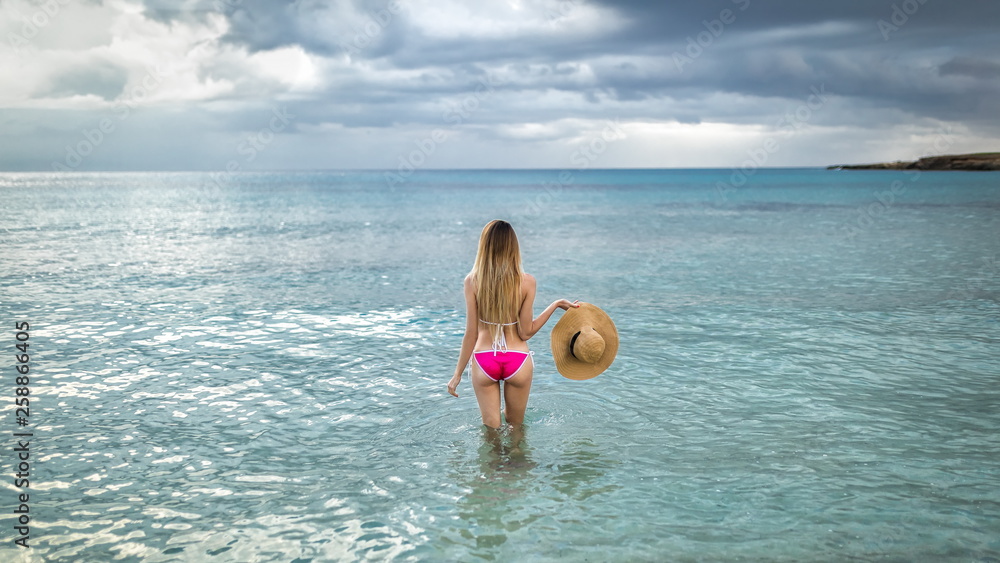 Blonde girl stands in water at sunset in pink bathing suit with summer hat in hand. Cloudy weather