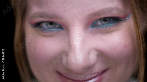 Close-up portrait of fair-haired model with colorful make-up joyfully moving her eyes on blurred lights background.