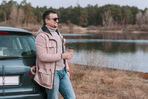 Young man driver stopped and resting with cup of coffee near car during travel break.