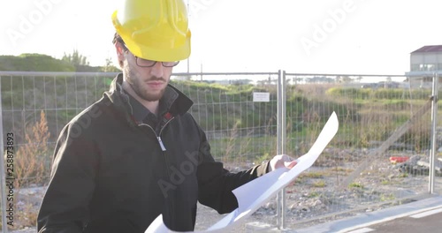 Giovanissimo ingegnere edile con caschetto giallo, occhiali e barba sta osservando la realizzazione dei lavori sul suo progetto tenuto in mano. photo