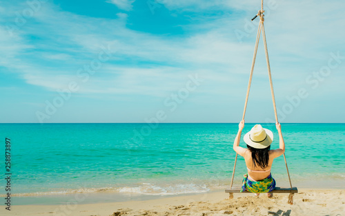Fototapeta Naklejka Na Ścianę i Meble -  Asian woman wear swimwear and hat swing the swings at sand beach  and looking beautiful tropical paradise sea and sky on sunny day. Summer vacation. Summer vibes. Enjoying and relaxing girl on holiday