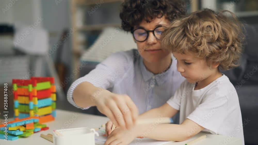 Adorable small child is drawing picture with pencils while proud mother is talking and smiling enjoying her son's talent. Childhood and parenthood concept. Adorable small child is drawing picture with pencils while proud mother is talking and smiling enjoying her son's talent. Childhood and parenthood concept.