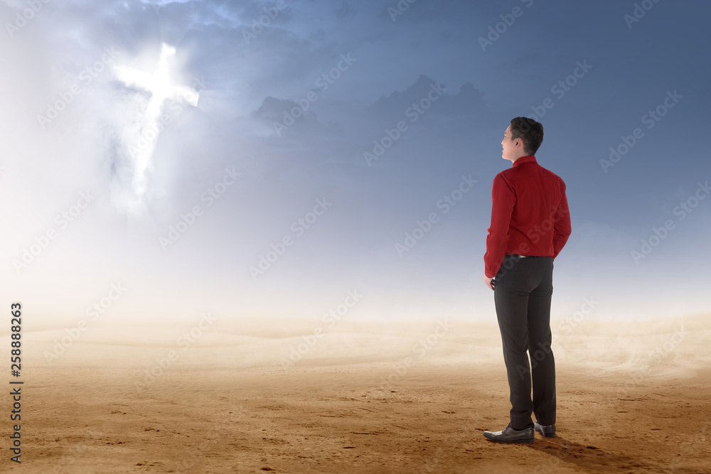 Rear view of asian businessman standing on desert and looking at glowing christian cross