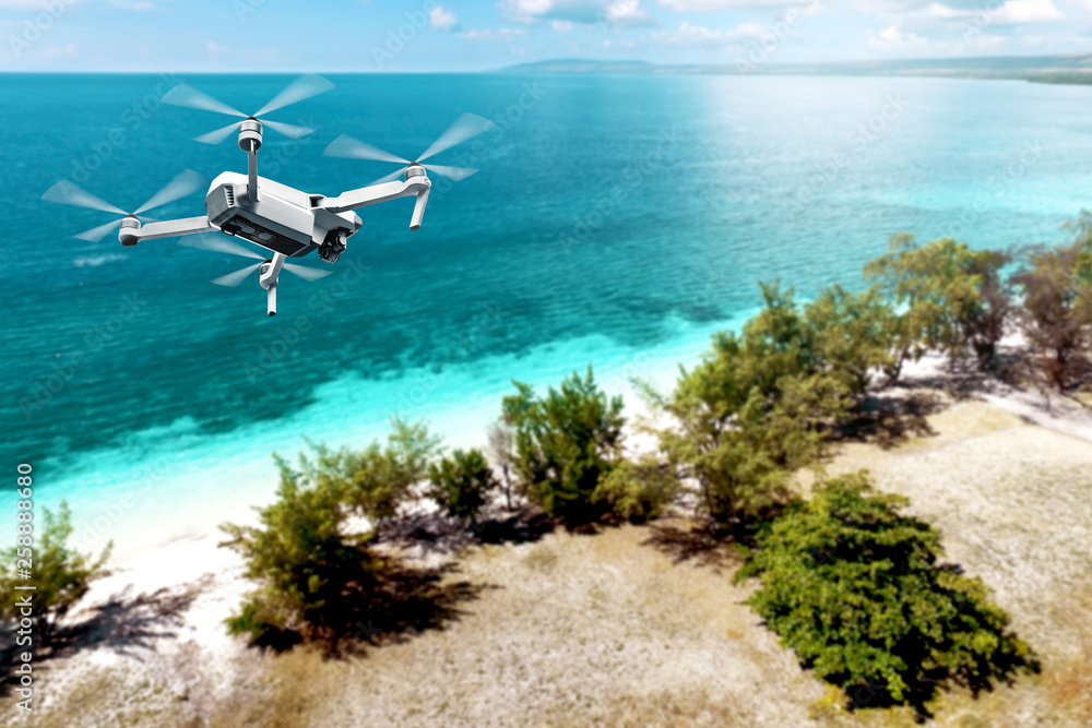 White drone with camera flying over the beach with green trees and blue ocean