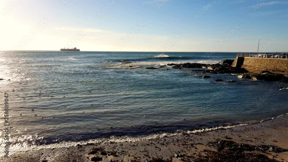 Ocean waters moving slow along shoreline in foreground with stone pier to right. Commercial ship passing along the horizon in the distance. Ocean waters moving slow along shoreline in foreground with stone pier to right. Commercial ship passing along the horizon in the distance.