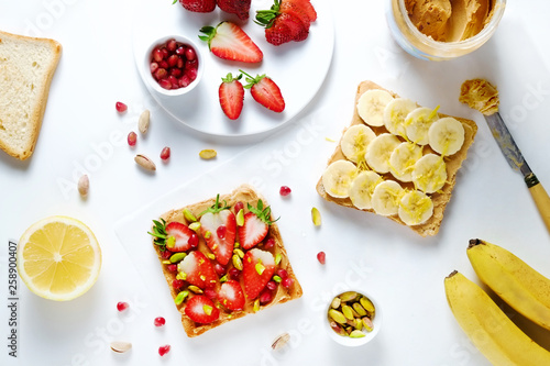 Healthy vegan breakfast concept. Peanut butter sandwiches with strawberry & banana, toasted bread, pomegranate seeds, pistachio nut. White table background. Top view, close up, copy space, flat lay