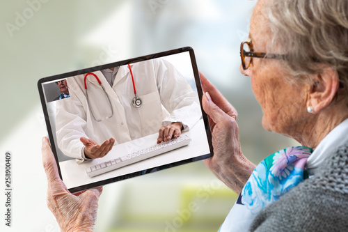 Senior woman consults a e-health doctor with tablet computer sitting in soft chair. In touchscreen, male doctor: With telehealth application patient can reach relevant specialist remotely. photo