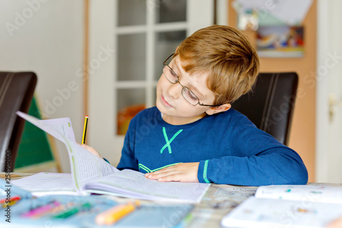 Portrait of cute school kid boy wearing glasses at home making homework. Little concentrated child writing with colorful pencils, indoors. Elementary school and education