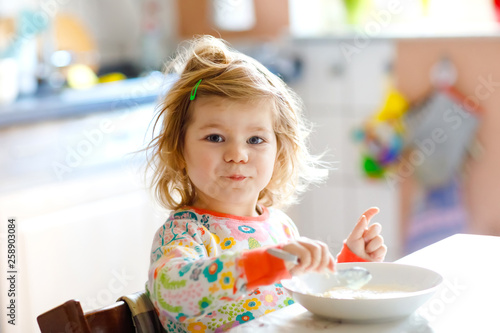 Adorable toddler girl eating healthy porrige from spoon for breakfast. Cute happy baby child in colorful pajamas sitting in kitchen and learning using spoon.