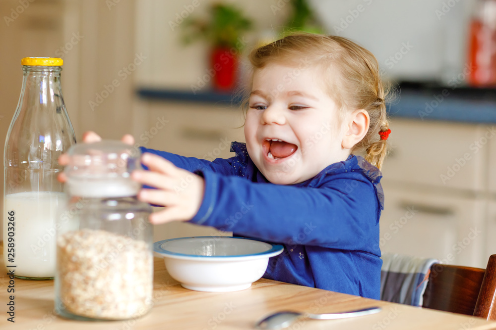 Adorable toddler girl eating healthy oatmeals with milk for breakfast. Cute happy baby child in colorful clothes sitting in kitchen and having fun with preparing oats, cereals. Indoors at home Adorable toddler girl eating healthy oatmeals with milk for breakfast. Cute happy baby child in colorful clothes sitting in kitchen and having fun with preparing oats, cereals. Indoors at home