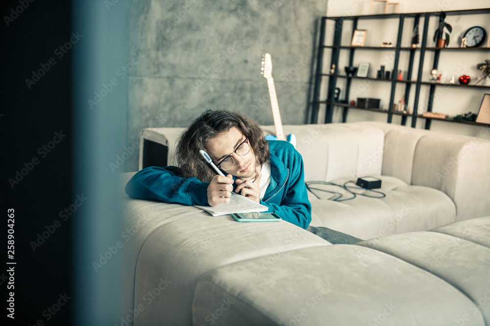 Naklejka premium Tired young dark-haired musician leaning on the couch and writing down