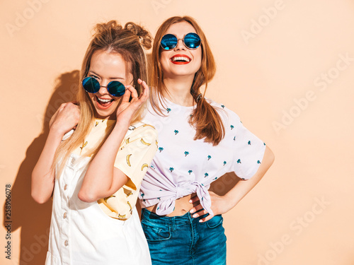 Two young beautiful smiling blond hipster girls in trendy summer colorful T-shirt clothes. Sexy carefree women posing near beige wall in round sunglasses. Positive models having fun © halayalex