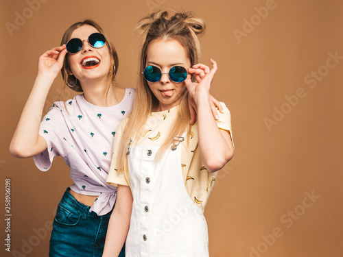 Two young beautiful smiling blond hipster girls in trendy summer colorful T-shirt clothes. Sexy carefree women posing near beige wall in round sunglasses. Positive models having fun © halayalex