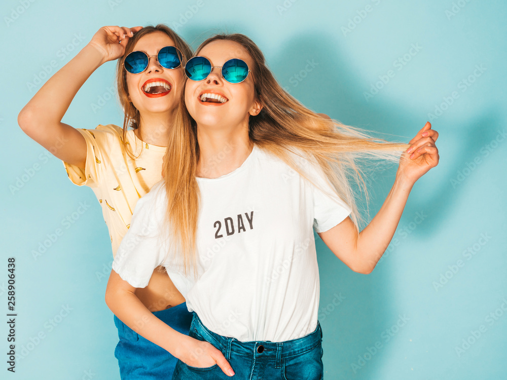 Two young beautiful smiling blond hipster girls in trendy summer jeans skirts clothes. Sexy carefree women posing near blue wall in round sunglasses. Positive models having fun and hugging Two young beautiful smiling blond hipster girls in trendy summer jeans skirts clothes. Sexy carefree women posing near blue wall in round sunglasses. Positive models having fun and hugging