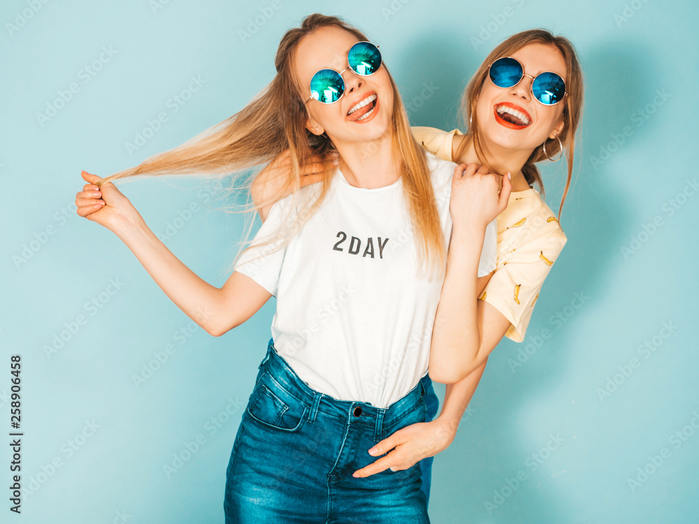 Two young beautiful smiling blond hipster girls in trendy summer jeans skirts clothes. Sexy carefree women posing near blue wall in round sunglasses. Positive models having fun and hugging Two young beautiful smiling blond hipster girls in trendy summer jeans skirts clothes. Sexy carefree women posing near blue wall in round sunglasses. Positive models having fun and hugging