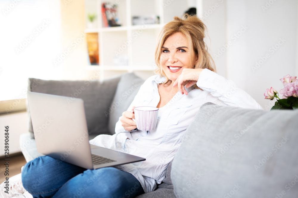 Sympathisch lächelnde Frau zuhause auf der Couch mit Laptop und Kaffee Sympathisch lächelnde Frau zuhause auf der Couch mit Laptop und Kaffee