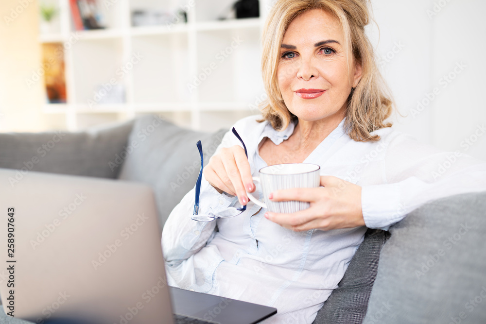 Attraktive reife Frau sitzt zuhause mit Kaffe und Latop auf der Couch Attraktive reife Frau sitzt zuhause mit Kaffe und Latop auf der Couch
